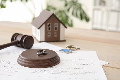 A model house, a gavel, a block with wedding rings on it, a set of keys and sheets of paper on a table.